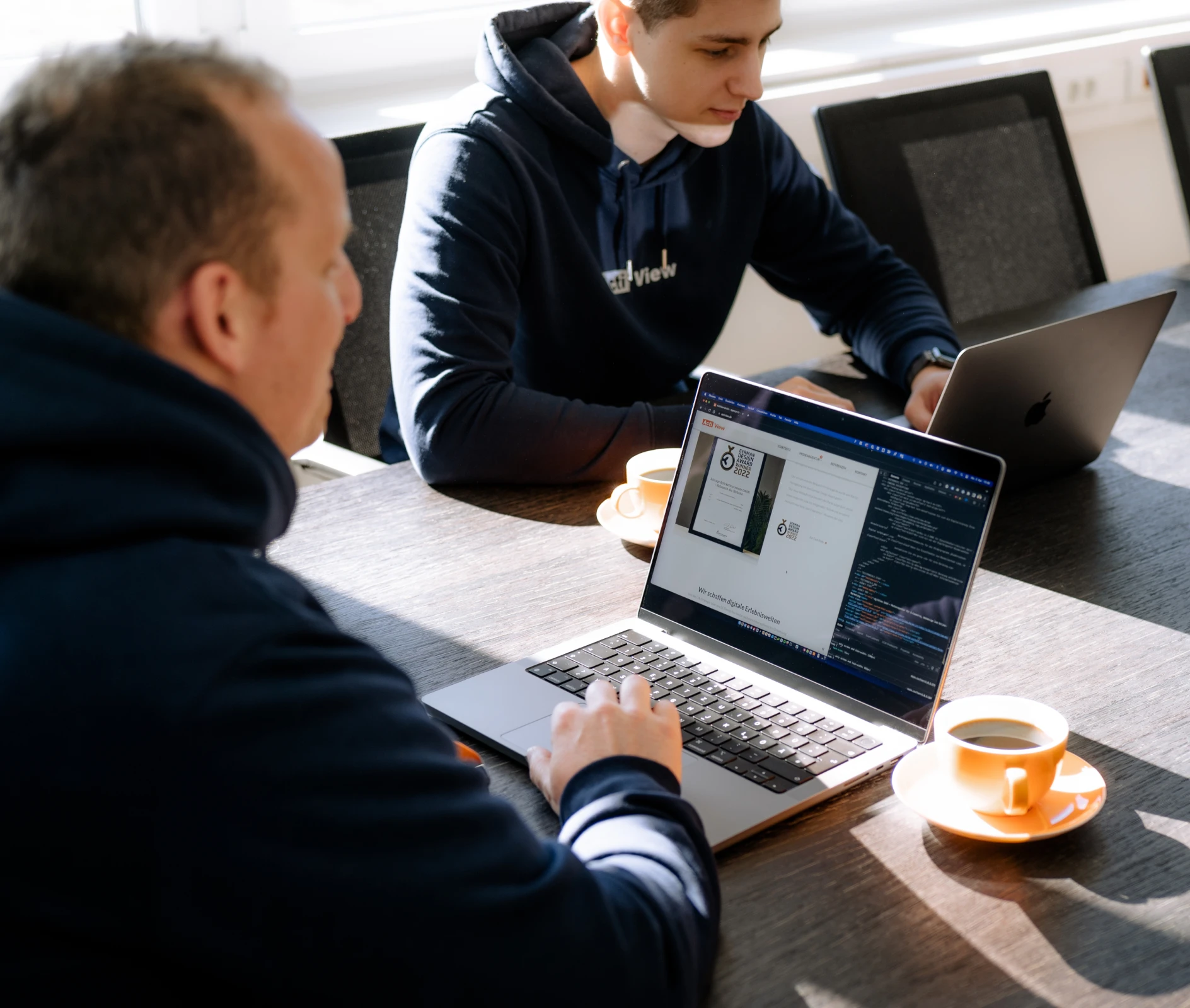 Zwei Personen in einem Business-Meeting, die auf einen Laptop mit Programmiercode schauen, mit einer Tasse Kaffee auf dem Tisch